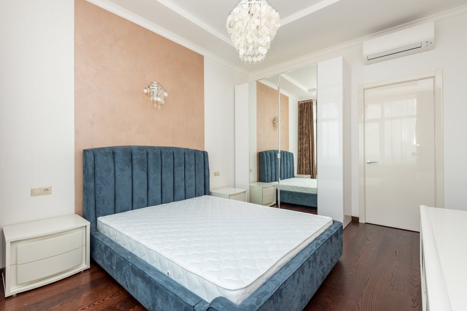 A tidy bedroom featuring a large, upholstered blue velvet bed with a white mattress, flanked by white bedside tables. The room has wooden flooring and a soft pink accent wall behind the bed, complemented by white walls and ceiling. There is a decorative chandelier hanging from the ceiling, and natural light filters through semi-transparent curtains in the background. A mirrored wardrobe with sliding doors reflects part of the room, enhancing its spacious feel. An air conditioning unit is mounted high on the wall near the door, which is beige. The overall appearance is clean and well-maintained, illustrating the high standards of surface cleaning and sanitisation promoted by Carpet Cleaning Belgravia.