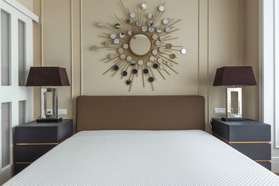 A neatly made bed with a beige upholstered headboard in a modern bedroom, featuring beige walls with minimal decorative molding, two black bedside tables with gold accents, each topped with a tall table lamp with a dark brown shade, and a round, sunburst-style mirror with multiple small circular mirrors and wooden rays mounted above the headboard. The room is illuminated by natural light from a large window on the right side, casting a bright and clean appearance suitable for surface cleaning and hygiene maintenance by Carpet Cleaning Belgravia, as part of their domestic cleaning and sanitisation services.