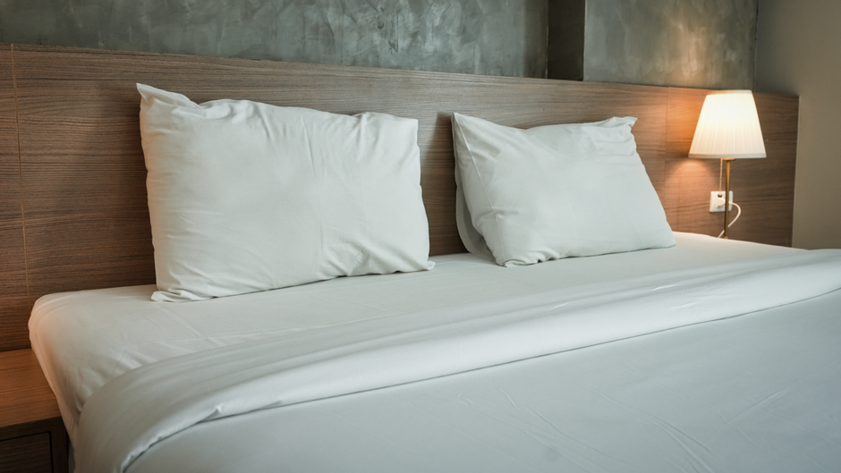 A tidy bedroom featuring a large, upholstered blue velvet bed with a white mattress, flanked by white bedside tables. The room has wooden flooring and a soft pink accent wall behind the bed, complemented by white walls and ceiling. There is a decorative chandelier hanging from the ceiling, and natural light filters through semi-transparent curtains in the background. A mirrored wardrobe with sliding doors reflects part of the room, enhancing its spacious feel. An air conditioning unit is mounted high on the wall near the door, which is beige. The overall appearance is clean and well-maintained, illustrating the high standards of surface cleaning and sanitisation promoted by Carpet Cleaning Belgravia.